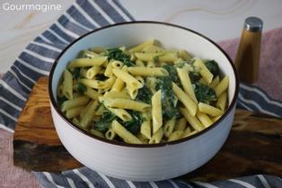 Eine weisse Schüssel gefüllt mit Penne, Krautstiel (Mangold) und Käse auf einem dunkelbraunen Holzbrett