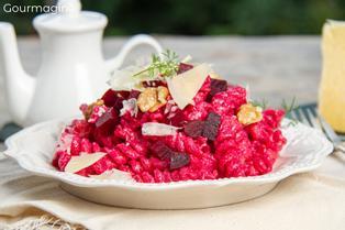 A white plate with pink pasta, beetroot pieces, cheese splinters and walnuts on a white tablecloth and a white teapot in the bacckground