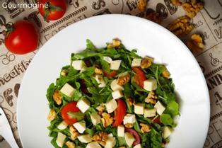 A white plate with green rocket salad, red cherry tomatoes, white mozzarella pieces and brown walnuts on a grey table cloth with tomatoes and walnuts in the background