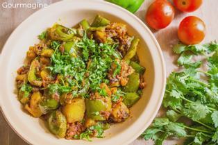 A white plate filled with fried green peppers, potatoes and onions on a curry sauce on a table with tomatoes and coriander