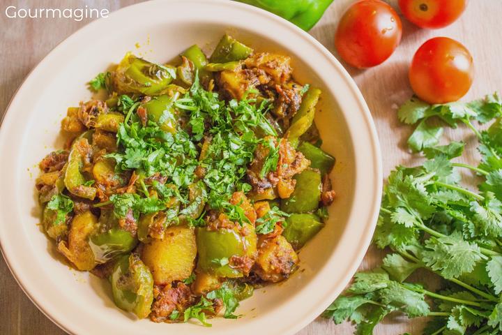 A white plate filled with fried green peppers, potatoes and onions on a curry sauce on a table with tomatoes and coriander