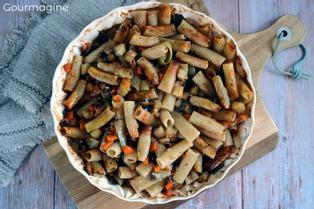 A white baking dish with baked penne and pieces of aubergines, carrots and peppers on a wooden board
